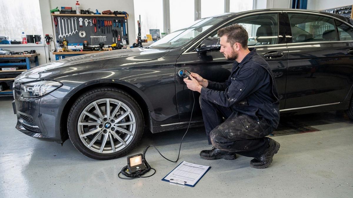 Independent inspector measuring paint depth along the door of a dark saloon car in a workshop bay